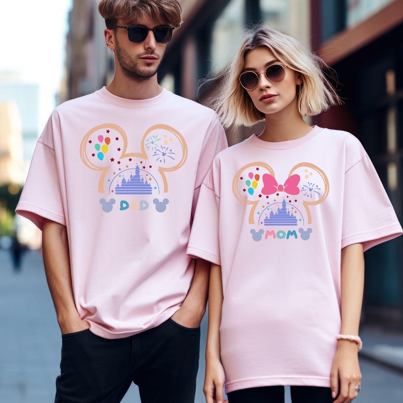 Pode incluir: Duas camisetas cor-de-rosa com um design de cabe&ccedil;a de Mickey Mouse que apresenta uma silhueta do castelo da Cinderela, bal&otilde;es e fogos de artif&iacute;cio. Os textos "DAD" e "MOM" est&atilde;o impressos nas camisetas.