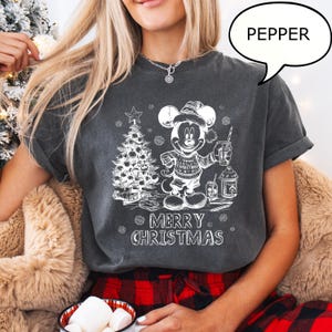 Puede incluir: Camiseta gris oscuro con un gr&aacute;fico blanco de Mickey Mouse con un su&eacute;ter navide&ntilde;o, sosteniendo una bebida y junto a un &aacute;rbol de Navidad. El texto "MERRY CHRISTMAS" est&aacute; debajo. Una burbuja de di&aacute;logo dice "PEPPER".