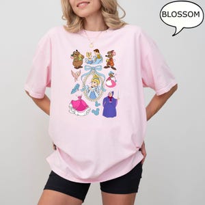 Puede incluir: Una camiseta rosa claro con un gr&aacute;fico colorido de Cenicienta y otros personajes de la pel&iacute;cula animada. El gr&aacute;fico incluye a Cenicienta, ratones, p&aacute;jaros y el hada madrina. La palabra "BLOSSOM" est&aacute; en una burbuja.