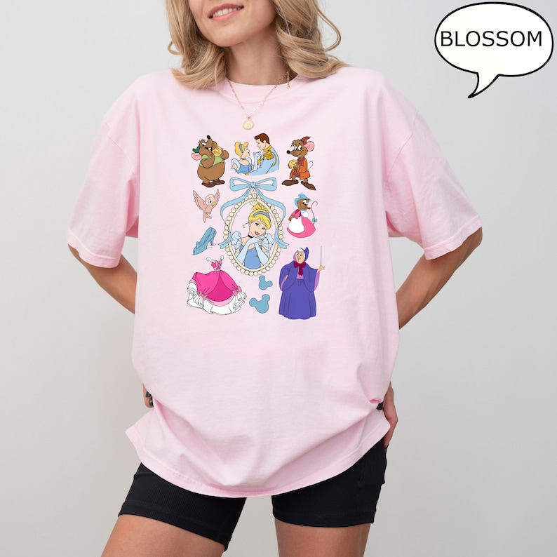 Puede incluir: Una camiseta rosa claro con un gr&aacute;fico colorido de Cenicienta y otros personajes de la pel&iacute;cula animada. El gr&aacute;fico incluye a Cenicienta, ratones, p&aacute;jaros y el hada madrina. La palabra "BLOSSOM" est&aacute; en una burbuja.