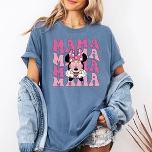 Puede incluir: Una camiseta azul con un gr&aacute;fico rosa y blanco de Minnie Mouse con el texto "MAMA" repetido en un patr&oacute;n apilado.