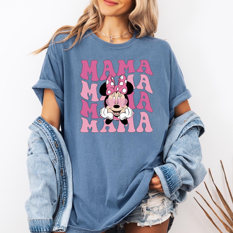 Puede incluir: Una camiseta azul con un gr&aacute;fico rosa y blanco de Minnie Mouse con el texto "MAMA" repetido en un patr&oacute;n apilado.