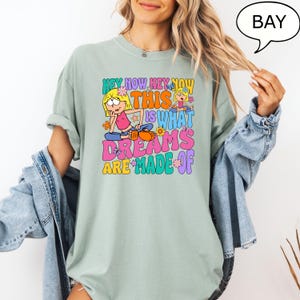 Może przedstawiać: Jasnozielony t-shirt z kolorowym nadrukiem graficznym przedstawiającym dziewczynkę z kresk&oacute;wki i tekst "Hey Now Hey Now This Is What Dreams Are Made Of".