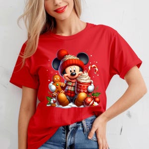 Puede incluir: Camiseta roja con un gr&aacute;fico navide&ntilde;o con Mickey Mouse con un gorro y una bufanda rojos y verdes, sosteniendo una taza de chocolate caliente y un hombre de jengibre.