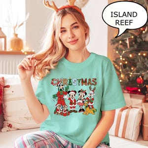 May include: A light teal t-shirt with a colourful Christmas graphic featuring Disney characters. The word "CHRISTMAS" is across the top. The image includes Mickey Mouse, Minnie Mouse, Goofy, and other characters in festive attire.