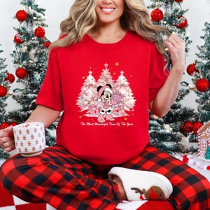 May include: A red t-shirt featuring a graphic of Minnie Mouse holding a gift in front of three Christmas trees. The text "The Most Wonderful Time Of The Year" is below the graphic. The person is wearing red and black plaid pajama pants.