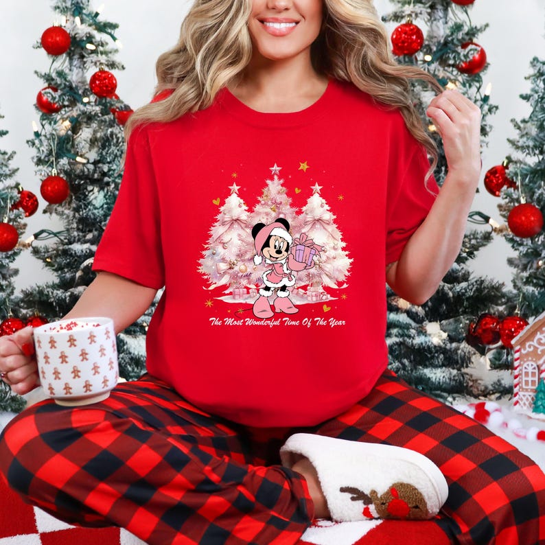 May include: A red t-shirt featuring a graphic of Minnie Mouse holding a gift in front of three Christmas trees. The text "The Most Wonderful Time Of The Year" is below the graphic. The person is wearing red and black plaid pajama pants.
