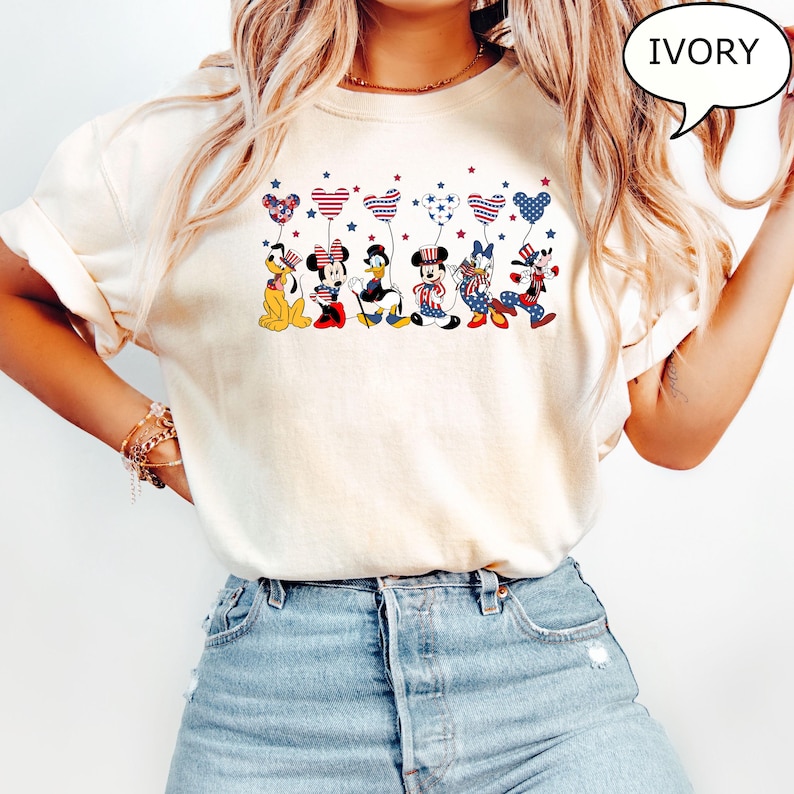 Puede incluir: Camiseta de color marfil con un dise&ntilde;o patri&oacute;tico con personajes de dibujos animados sosteniendo globos en forma de coraz&oacute;n. Los globos est&aacute;n decorados con estrellas y rayas rojas, blancas y azules. La camiseta tiene la palabra "IVORY" en una burbuja.