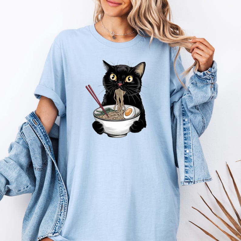 Puede incluir: Una camiseta azul claro con un gato negro comiendo fideos ramen de un cuenco blanco. El gato tiene ojos verdes y sostiene palillos en sus patas.