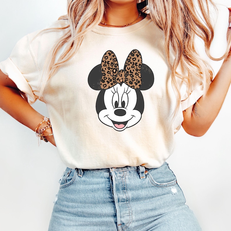 May include: A white t-shirt with a black and white cartoon image of Minnie Mouse. Minnie Mouse is wearing a brown and black leopard print bow.