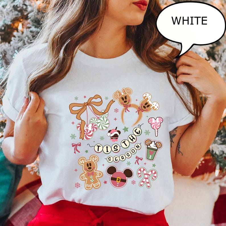 May include: White t-shirt with a festive design featuring gingerbread men, candy canes, and the phrase "Tis the Season." The design includes a Mickey Mouse-inspired gingerbread cookie and a cup of hot cocoa.