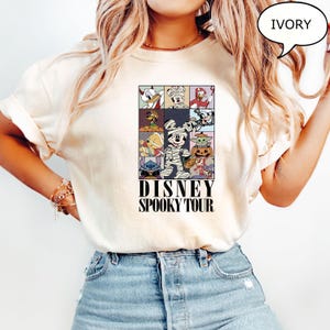 May include: Ivory-colored t-shirt featuring a colorful graphic of Disney characters in Halloween costumes. The design includes Donald Duck, Mickey Mouse, and Stitch. The text "DISNEY SPOOKY TOUR" is printed below the graphic.