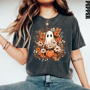 Op de afbeelding: Een donkergrijs T-shirt met een witte geest omringd door oranje en witte bloemen en een kleine oranje pompoen. De tekst "Pepper" staat in de rechterbovenhoek van de afbeelding.