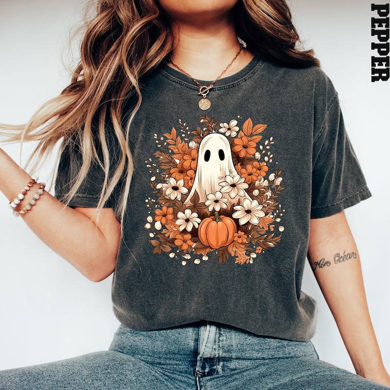 Op de afbeelding: Een donkergrijs T-shirt met een witte geest omringd door oranje en witte bloemen en een kleine oranje pompoen. De tekst "Pepper" staat in de rechterbovenhoek van de afbeelding.