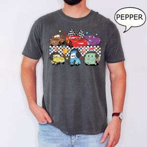 May include: A dark gray t-shirt with a checkered pattern and a graphic of cartoon cars from the movie Cars. The cars are in a line and include Lightning McQueen, Mater, and others. The text "PEPPER" is in a speech bubble in the upper right corner of the image.