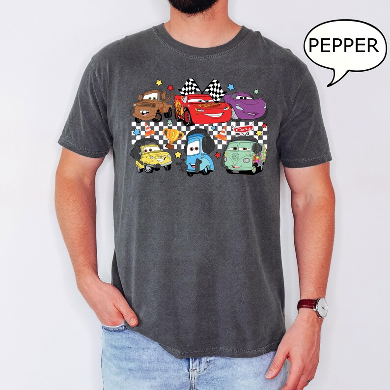 May include: A dark gray t-shirt with a checkered pattern and a graphic of cartoon cars from the movie Cars. The cars are in a line and include Lightning McQueen, Mater, and others. The text "PEPPER" is in a speech bubble in the upper right corner of the image.