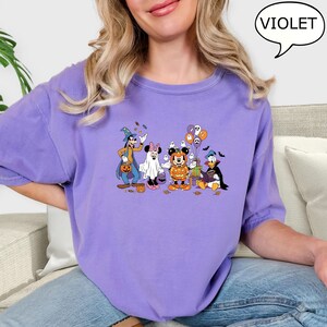 May include: A violet-colored t-shirt featuring a Halloween-themed graphic of Disney characters, including Goofy, Minnie Mouse, Mickey Mouse, and Donald Duck, in costumes. The word "VIOLET" is in a speech bubble.