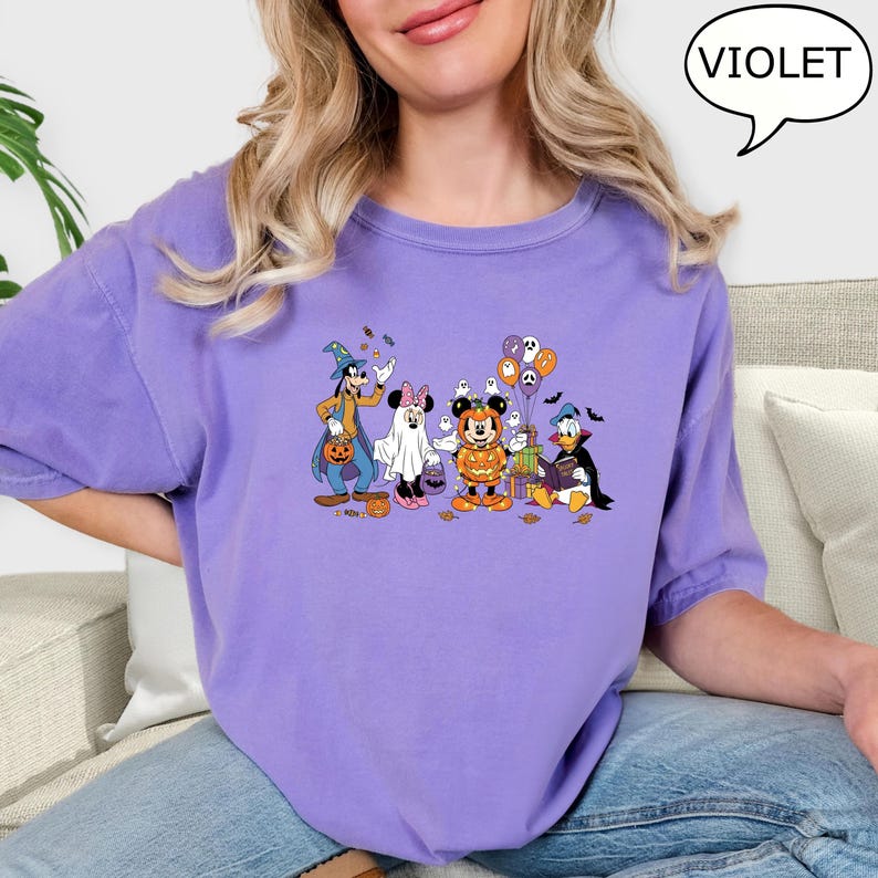 May include: A violet-colored t-shirt featuring a Halloween-themed graphic of Disney characters, including Goofy, Minnie Mouse, Mickey Mouse, and Donald Duck, in costumes. The word "VIOLET" is in a speech bubble.