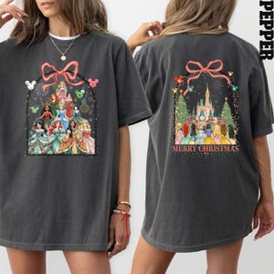 May include: Dark grey t-shirt with a festive design featuring Disney princesses, fireworks, and a pink bow. The back of the shirt shows a castle, Christmas trees, and the text "MERRY CHRISTMAS".