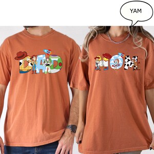 May include: Two orange t-shirts, one with the word "DAD" and the other with "MOM", in a colourful, cartoon style. The "DAD" shirt features characters from a popular animated film. The "MOM" shirt has similar design elements.