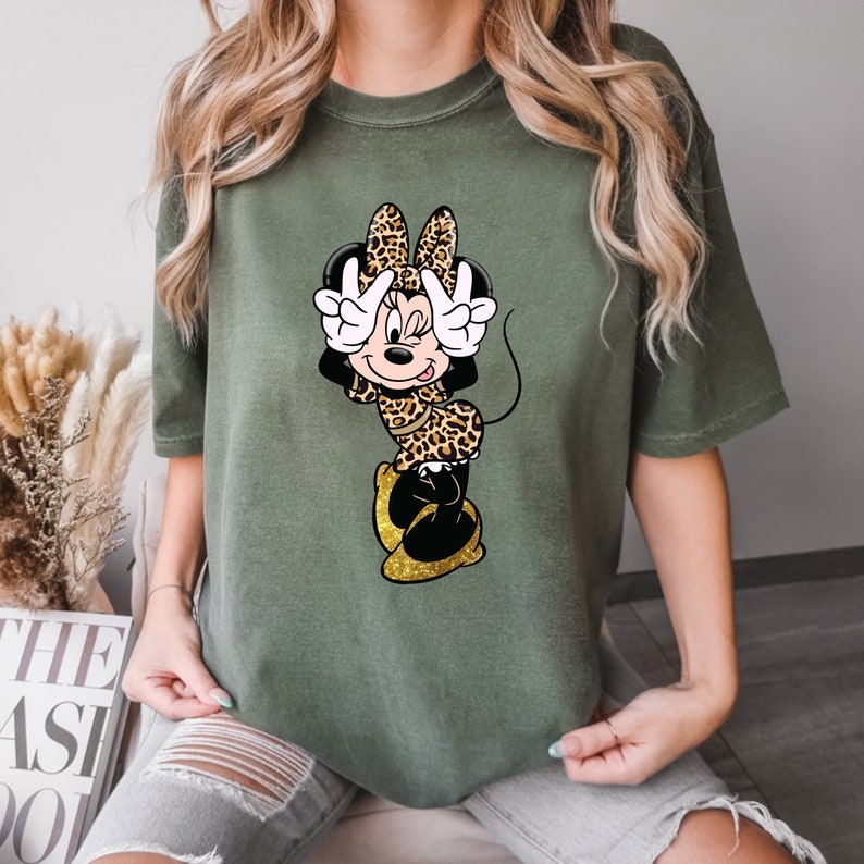 K&ouml;nnte beinhalten: Olivgr&uuml;nes T-Shirt mit einer Cartoon-Minnie-Maus in einem Leopardenmuster-Outfit. Minnie macht mit beiden H&auml;nden ein Friedenszeichen. Ihre Schuhe sind goldfarben glitzernd. Das Shirt ist ein l&auml;ssiger, bequemer Stil.