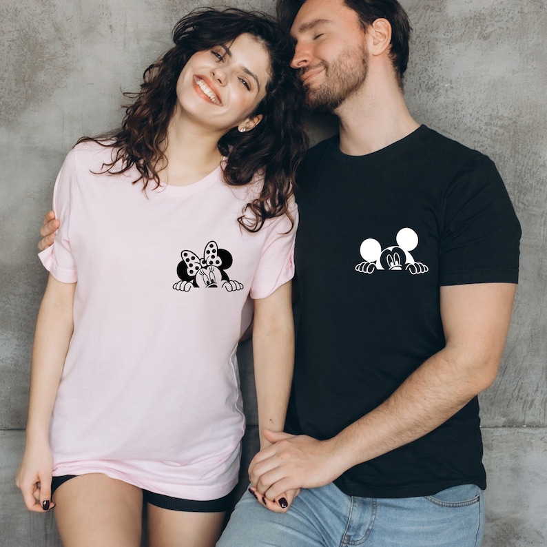 May include: A couple wearing matching t-shirts with a Mickey Mouse and Minnie Mouse design. The woman's shirt is pink and features Minnie Mouse peeking out from behind a heart. The man's shirt is black and features Mickey Mouse peeking out from behind a heart.