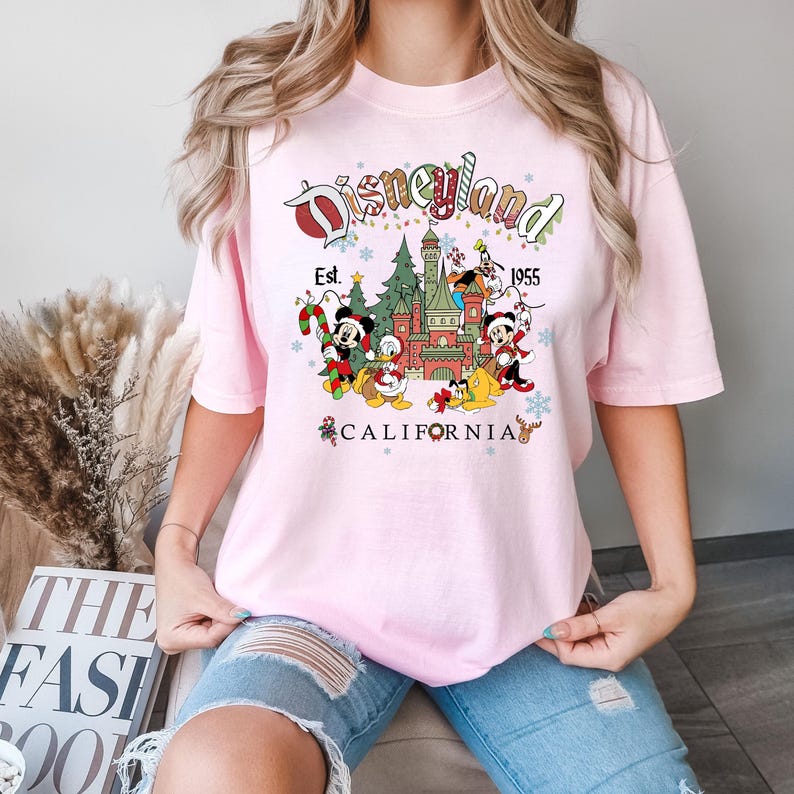 Puede incluir: Camiseta rosa con un gr&aacute;fico de Disneyland con tem&aacute;tica navide&ntilde;a. El gr&aacute;fico presenta a Mickey Mouse, Donald Duck, Goofy y Pluto frente al castillo de la Bella Durmiente. El texto dice "Disneyland Est. 1955 California".