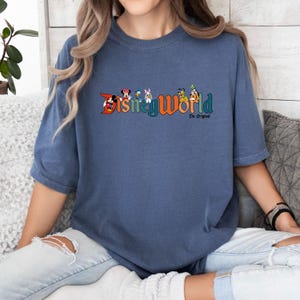 K&ouml;nnte beinhalten: Ein blaues T-Shirt mit einer Grafik im Vintage-Stil des Disney World-Logos und verschiedenen Disney-Charakteren. Der Text unter dem Logo lautet "The Original".