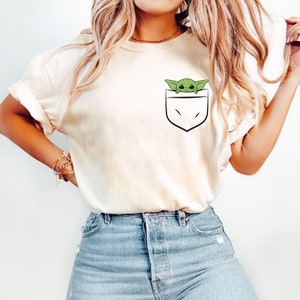 May include: A white t-shirt with a green character peeking out of a pocket on the front. The character is from the Star Wars franchise.