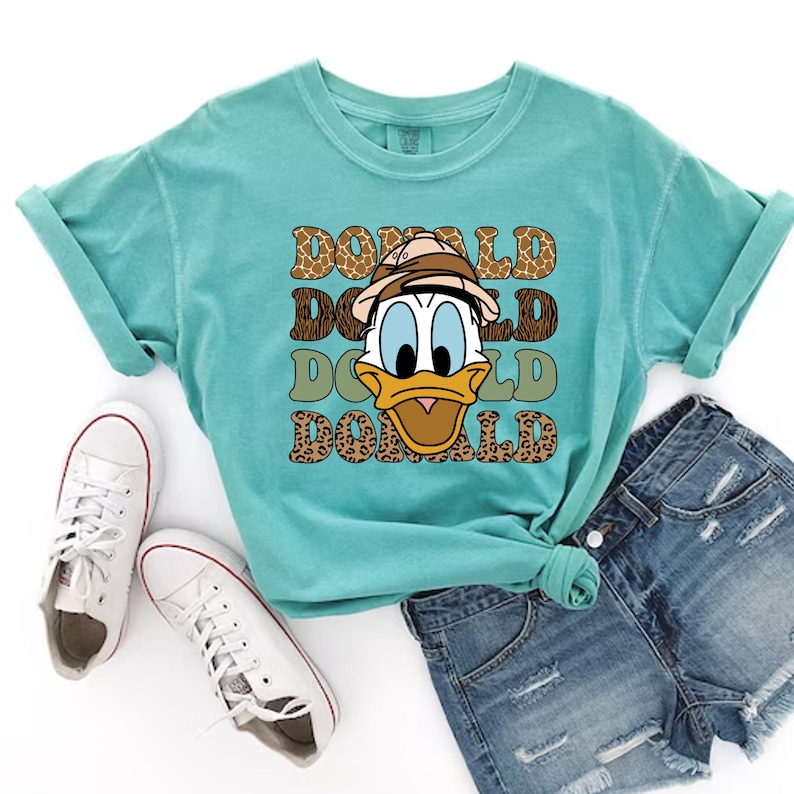 May include: A turquoise blue t-shirt featuring a cartoon image of Donald Duck wearing a safari hat. The text "DONALD" is repeated four times in a retro font, with each word layered on top of a different animal print.