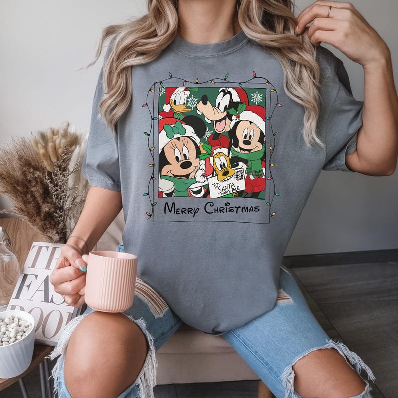 May include: A gray t-shirt with a Christmas-themed graphic featuring Mickey Mouse, Minnie Mouse, Goofy, and Donald Duck. The graphic is framed in a Polaroid-style frame with string lights around it. The text "Merry Christmas" is printed below the graphic.