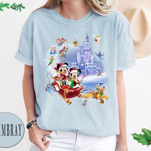 Puede incluir: Una camiseta azul claro con un dise&ntilde;o navide&ntilde;o que presenta a Mickey Mouse y Minnie Mouse en un trineo tirado por Pluto. El dise&ntilde;o tambi&eacute;n incluye el castillo de Cenicienta y otros personajes de Disney.