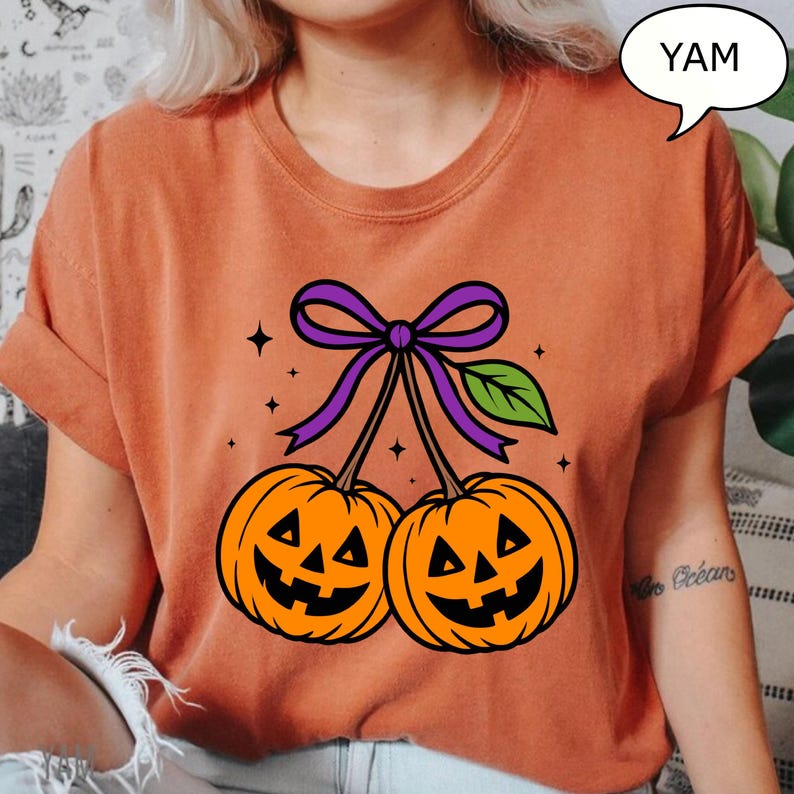 May include: Orange t-shirt featuring a Halloween-themed design. The graphic shows two orange jack-o'-lanterns with smiling faces, connected by stems and a purple bow. A green leaf and stars complete the design. The word "YAM" is in a speech bubble.