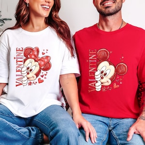 Puede incluir: Camisetas blancas y rojas de San Valent&iacute;n con un personaje de rat&oacute;n de dibujos animados con un lazo rojo y corazones. La palabra "VALENTINE" est&aacute; impresa verticalmente en el lado izquierdo del dise&ntilde;o. Las camisetas son usadas por dos personas.