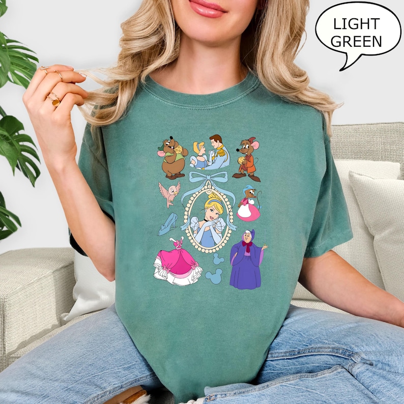 Puede incluir: Camiseta verde claro con un dise&ntilde;o tem&aacute;tico de Cenicienta. Las ilustraciones incluyen a Cenicienta, su hada madrina, ratones, p&aacute;jaros y otros personajes. El texto "LIGHT GREEN" est&aacute; en un bocadillo.