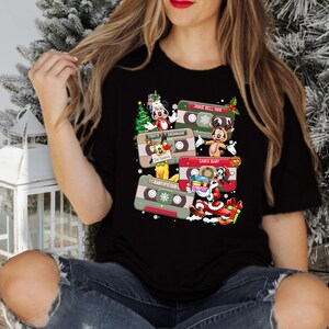 Puede incluir: Camiseta negra con un dise&ntilde;o navide&ntilde;o con personajes de dibujos animados y cintas de casete. El texto en las cintas incluye "Jingle Bell Rock", "Frosty", "Snowman", "Santa Baby", "Baby It's Cold Outside" y "'Tis the Season".