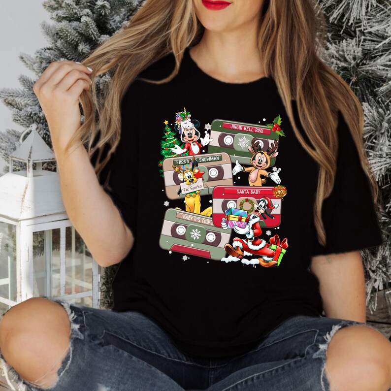 Puede incluir: Camiseta negra con un dise&ntilde;o navide&ntilde;o con personajes de dibujos animados y cintas de casete. El texto en las cintas incluye "Jingle Bell Rock", "Frosty", "Snowman", "Santa Baby", "Baby It's Cold Outside" y "'Tis the Season".