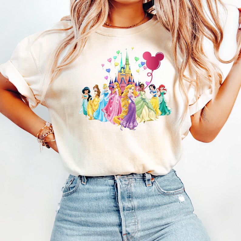 May include: A white t-shirt with a colorful graphic of Disney princesses standing in front of Cinderella's castle. The graphic also includes a pink Mickey Mouse balloon with hearts floating around the castle.