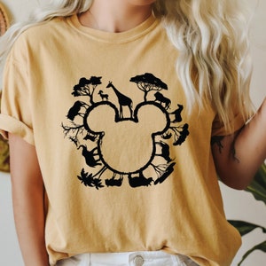 Puede incluir: Una camiseta amarilla con una silueta negra de una cabeza de Mickey Mouse rodeada de animales y &aacute;rboles. Los animales incluyen una jirafa, una cebra, un le&oacute;n y un elefante.