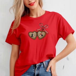 May include: Red t-shirt with a heart-shaped locket featuring Mickey and Minnie Mouse. The locket has a pink bow and is hanging from a chain.