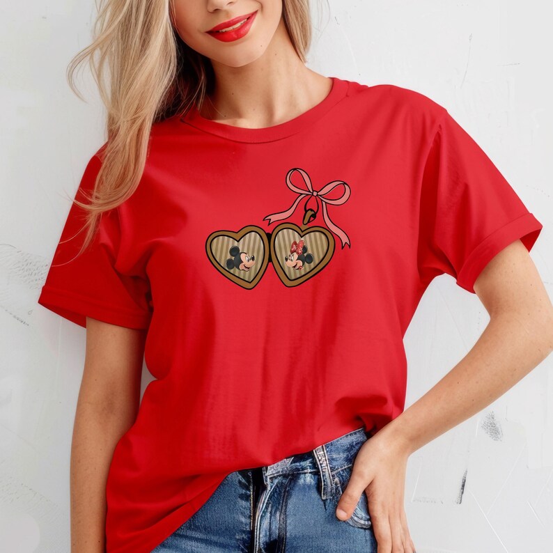 May include: Red t-shirt with a heart-shaped locket featuring Mickey and Minnie Mouse. The locket has a pink bow and is hanging from a chain.