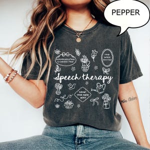 May include: Dark gray t-shirt with the words "Speech therapy" in a script font, surrounded by white line art illustrations of flowers, a watering can, and other designs. The text "Communication is Connection" is also visible.
