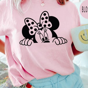 K&ouml;nnte beinhalten: Ein pinkfarbenes T-Shirt mit einer schwarz-wei&szlig;en Illustration von Minnie Mouse, die &uuml;ber den Rand schaut. Die Illustration enth&auml;lt Minnies charakteristisches gepunktetes Band.