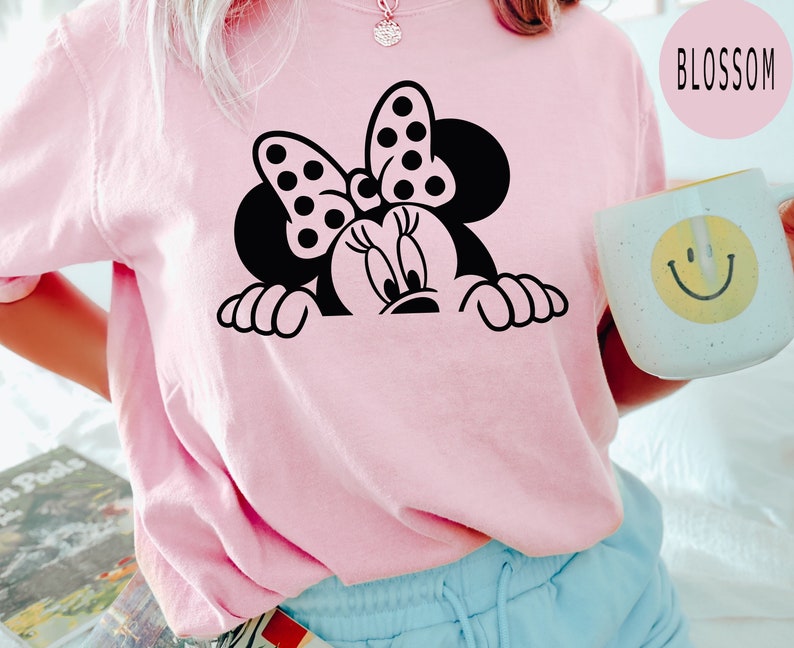 K&ouml;nnte beinhalten: Ein pinkfarbenes T-Shirt mit einer schwarz-wei&szlig;en Illustration von Minnie Mouse, die &uuml;ber den Rand schaut. Die Illustration enth&auml;lt Minnies charakteristisches gepunktetes Band.