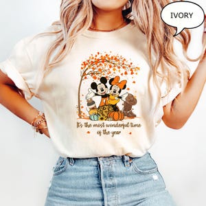 May include: Ivory-colored t-shirt featuring Mickey and Minnie Mouse under a tree with autumn leaves. The graphic includes pumpkins, a cowboy boot, and the text "It's the most wonderful time of the year."