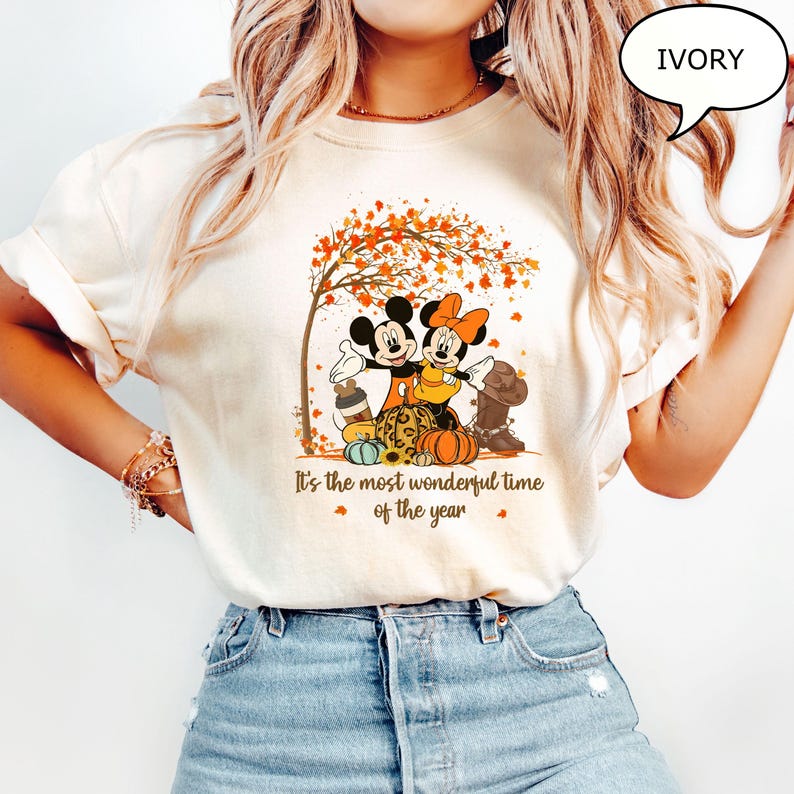 May include: Ivory-colored t-shirt featuring Mickey and Minnie Mouse under a tree with autumn leaves. The graphic includes pumpkins, a cowboy boot, and the text "It's the most wonderful time of the year."