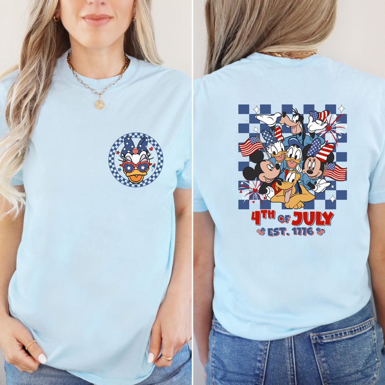 Pode incluir: Camiseta azul claro com a Pata Daisy usando &oacute;culos patri&oacute;ticos na frente. A parte de tr&aacute;s mostra Mickey Mouse, Donald Duck, Pateta e Pluto com bandeiras americanas e fogos de artif&iacute;cio, com o texto "4th of July" e "Est. 1776".