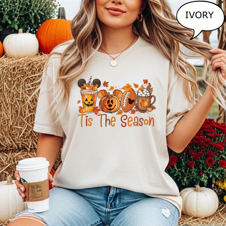 May include: Ivory-colored t-shirt featuring a fall-themed design. The design includes a pumpkin spice latte with a Mickey Mouse face, a Mickey Mouse pumpkin, a football with a Mickey Mouse face, and a mug of hot cocoa. The words 'Tis The Season' are printed in orange.