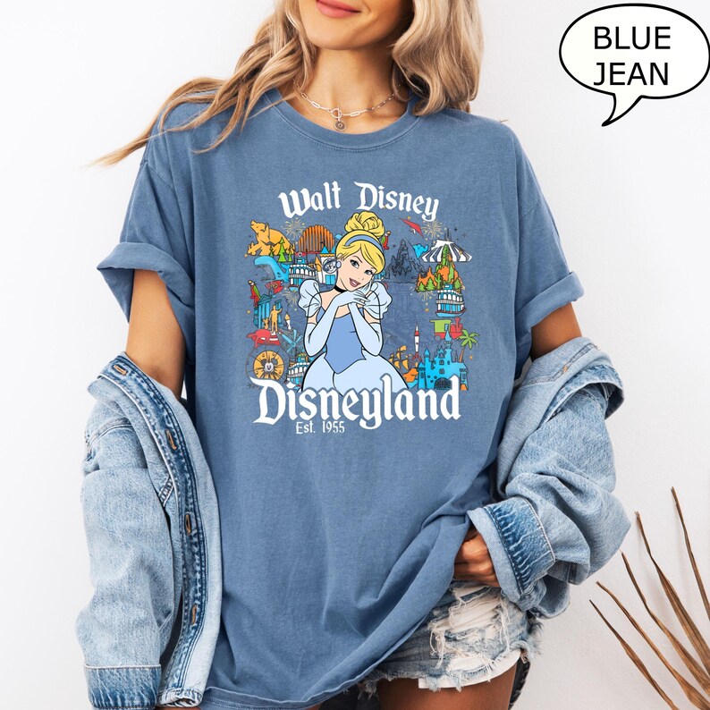 May include: A blue jean-coloured t-shirt with a Cinderella graphic and the text "Walt Disney Disneyland Est. 1955". The graphic includes Cinderella in a blue dress and various Disneyland attractions.