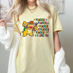 May include: A light yellow t-shirt featuring a cartoon Bowser character holding a microphone and singing. The text "PEACHES I LOVE YOU" is printed in colorful letters and musical notes.